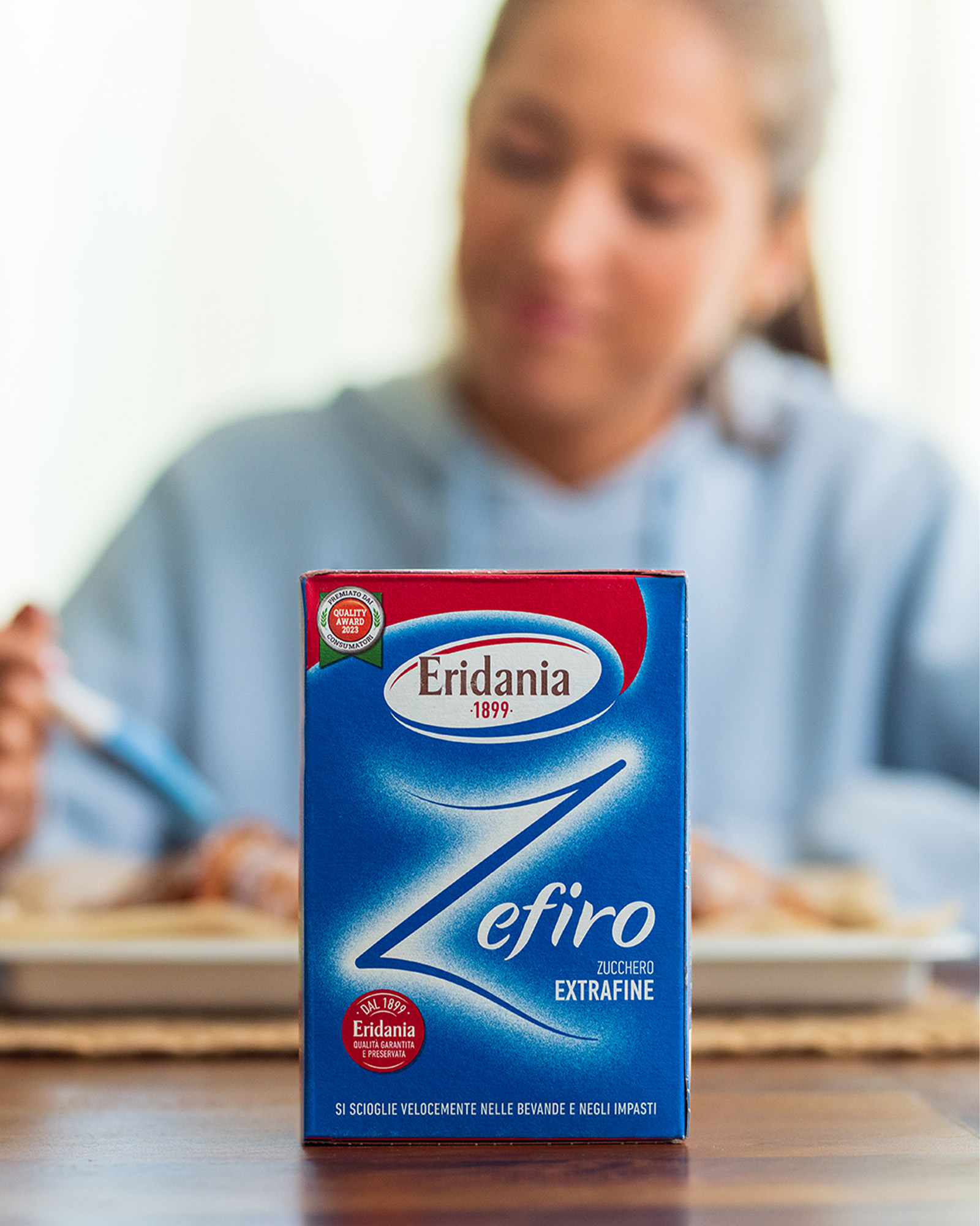 Confezione blu e rossa di zucchero extrafine eridania zefiro in primo piano su un tavolo sullo sfondo sfocato una donna seduta sta mangiando