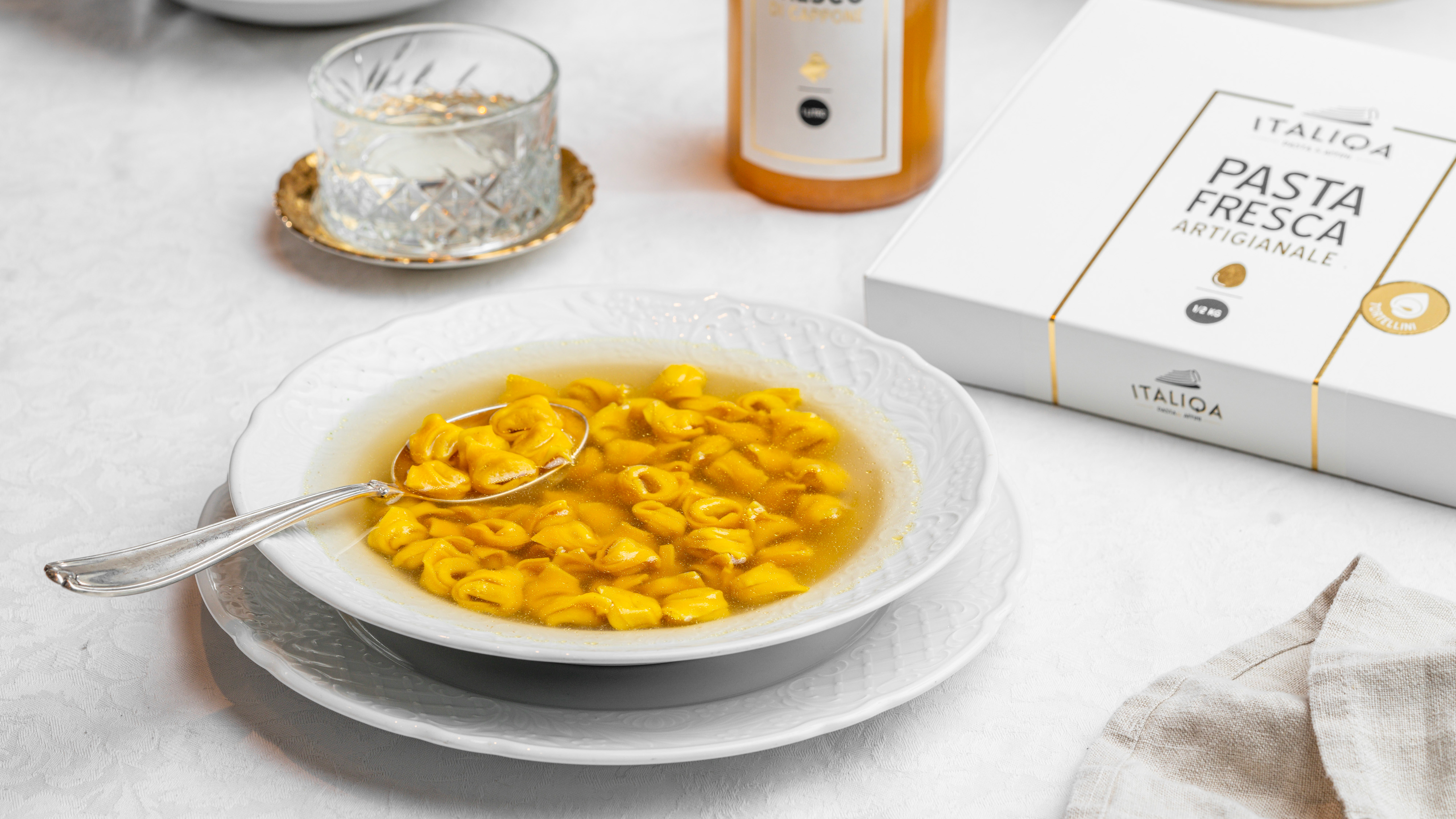 Piatto di tortellini in brodo accanto a confezione di pasta fresca artigianale italiqa bicchiere e bottiglia su tavola bianca