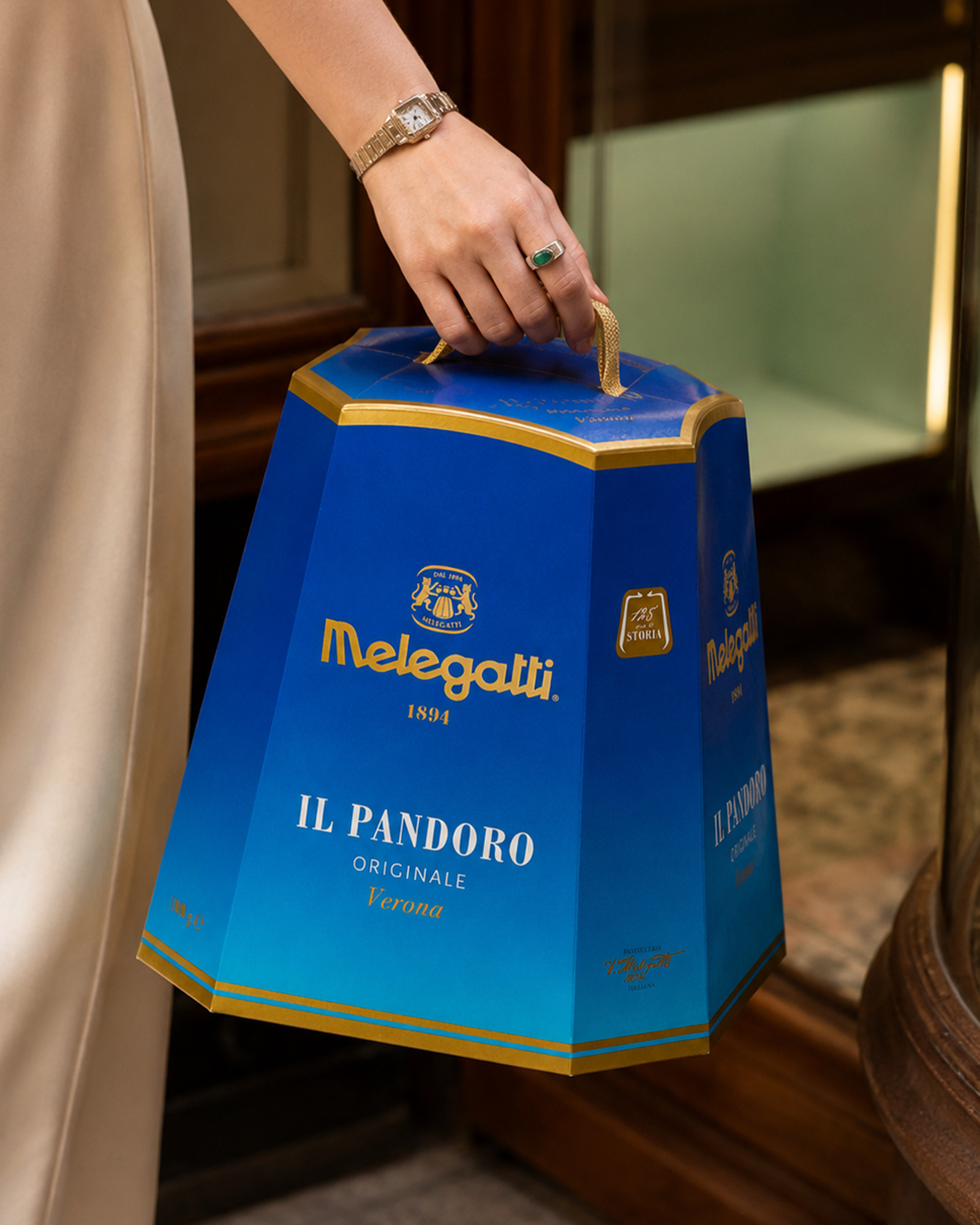 Persona con abito chiaro che trasporta una confezione blu e oro del pandoro originale melegatti tenendola per il manico