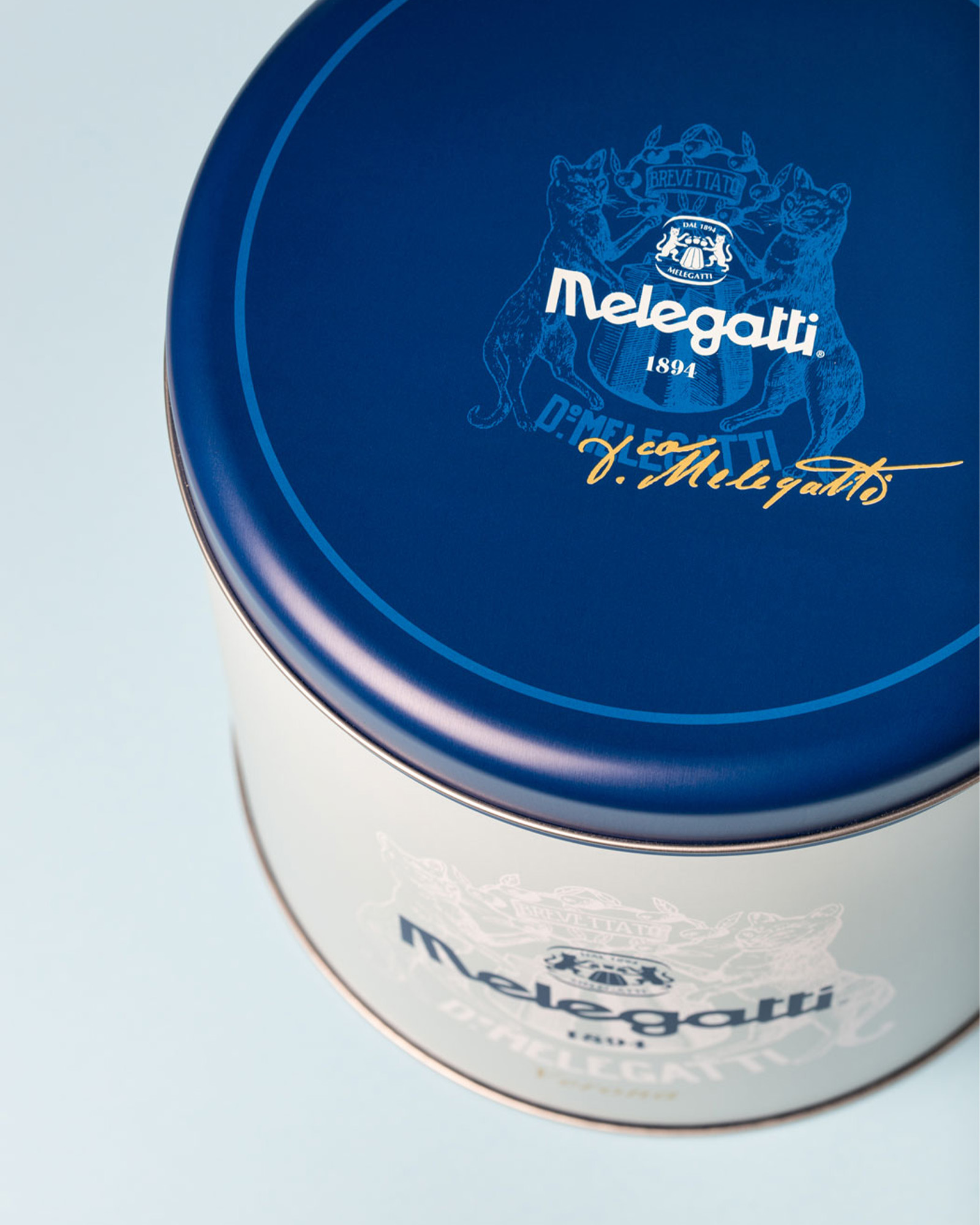 Contenitore metallico bianco con coperchio blu melegatti decorato con logo stemma e firma su sfondo chiaro
