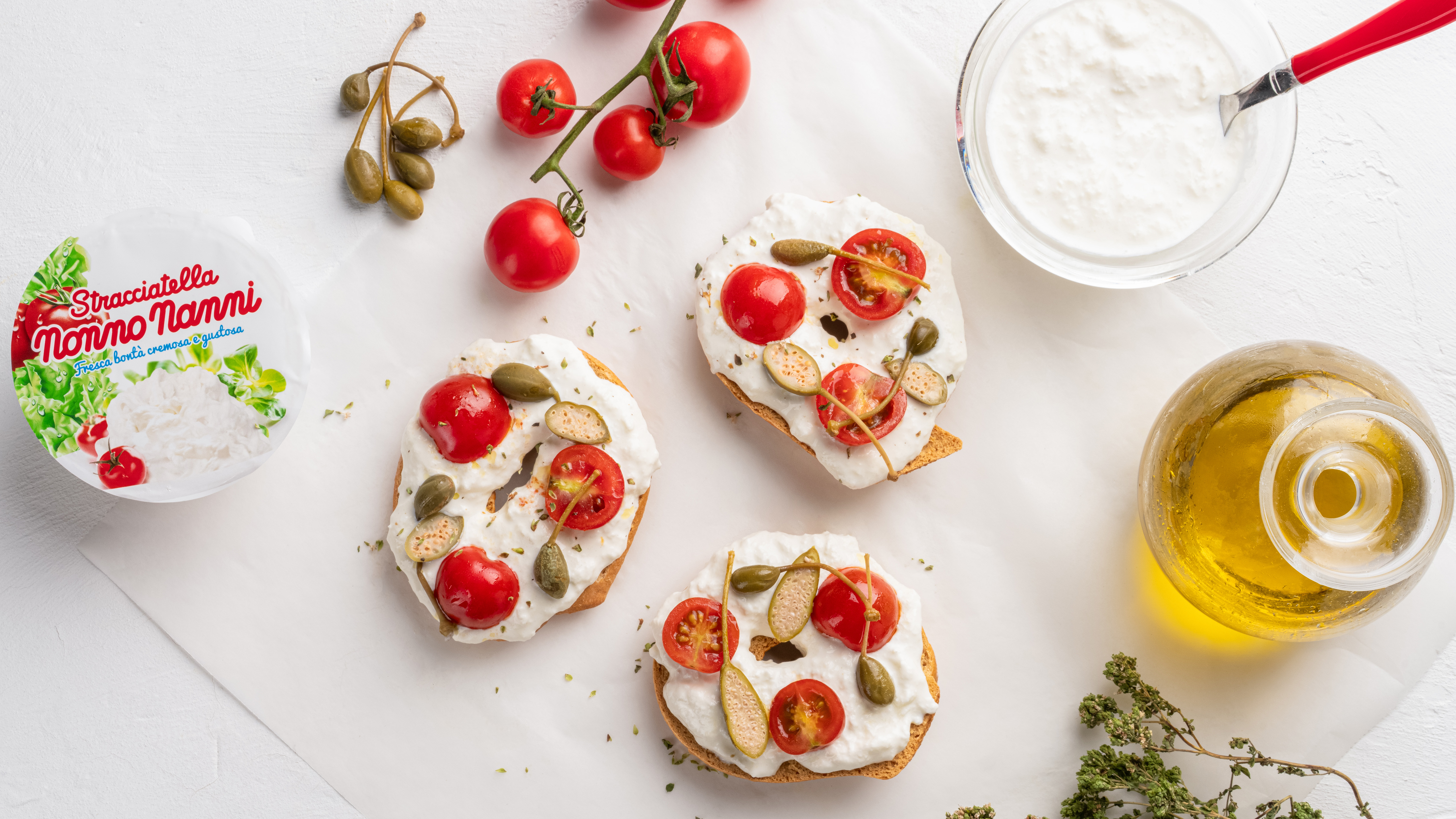 Bruschette con stracciatella pomodorini e capperi accanto a confezione nonno nanni ciotola di formaggio e bottiglia di olio su sfondo bianco