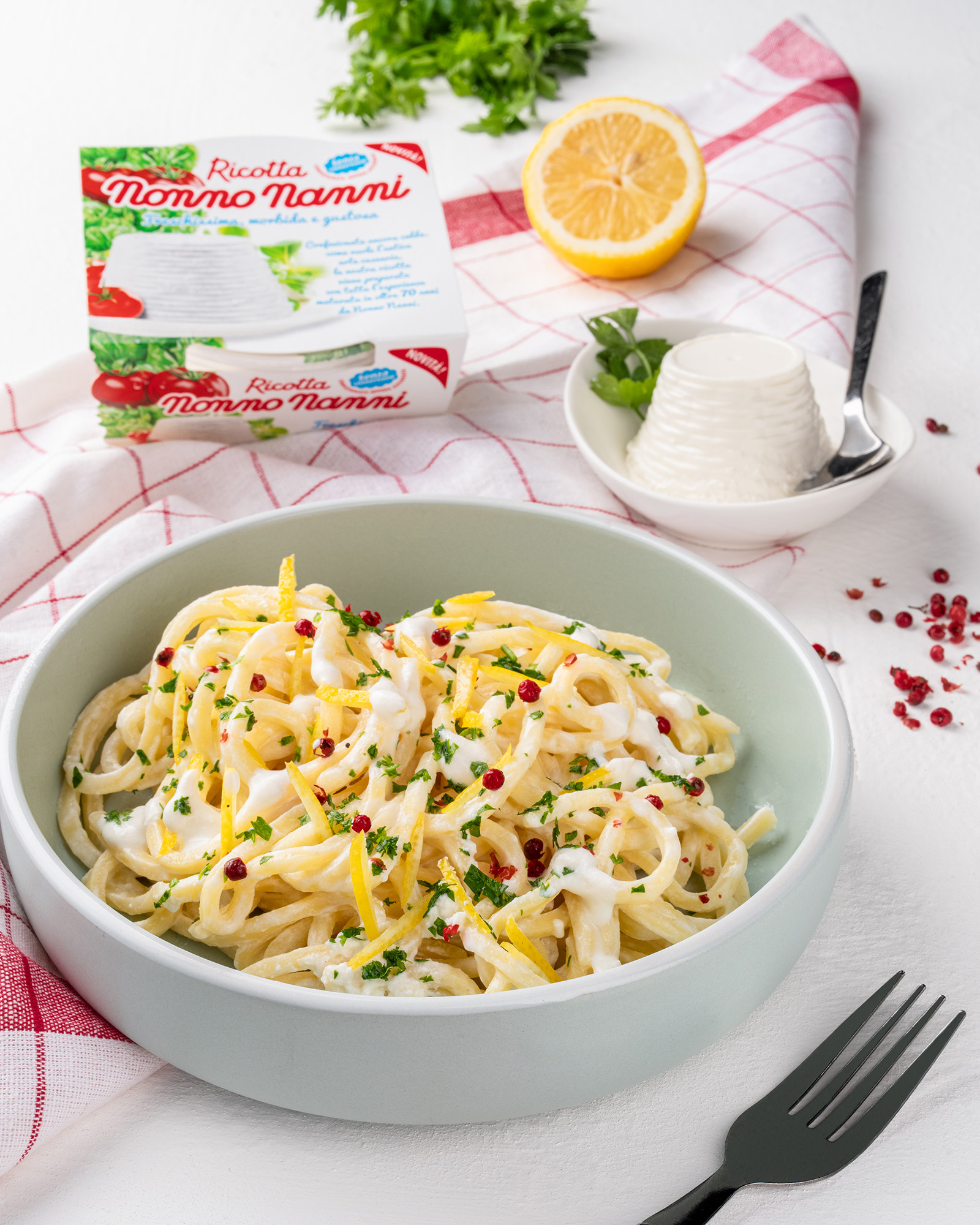 Piatto di spaghetti con crema di ricotta limone prezzemolo e pepe rosa accanto a confezione di ricotta nonno nanni e ciotola di ricotta su sfondo bianco