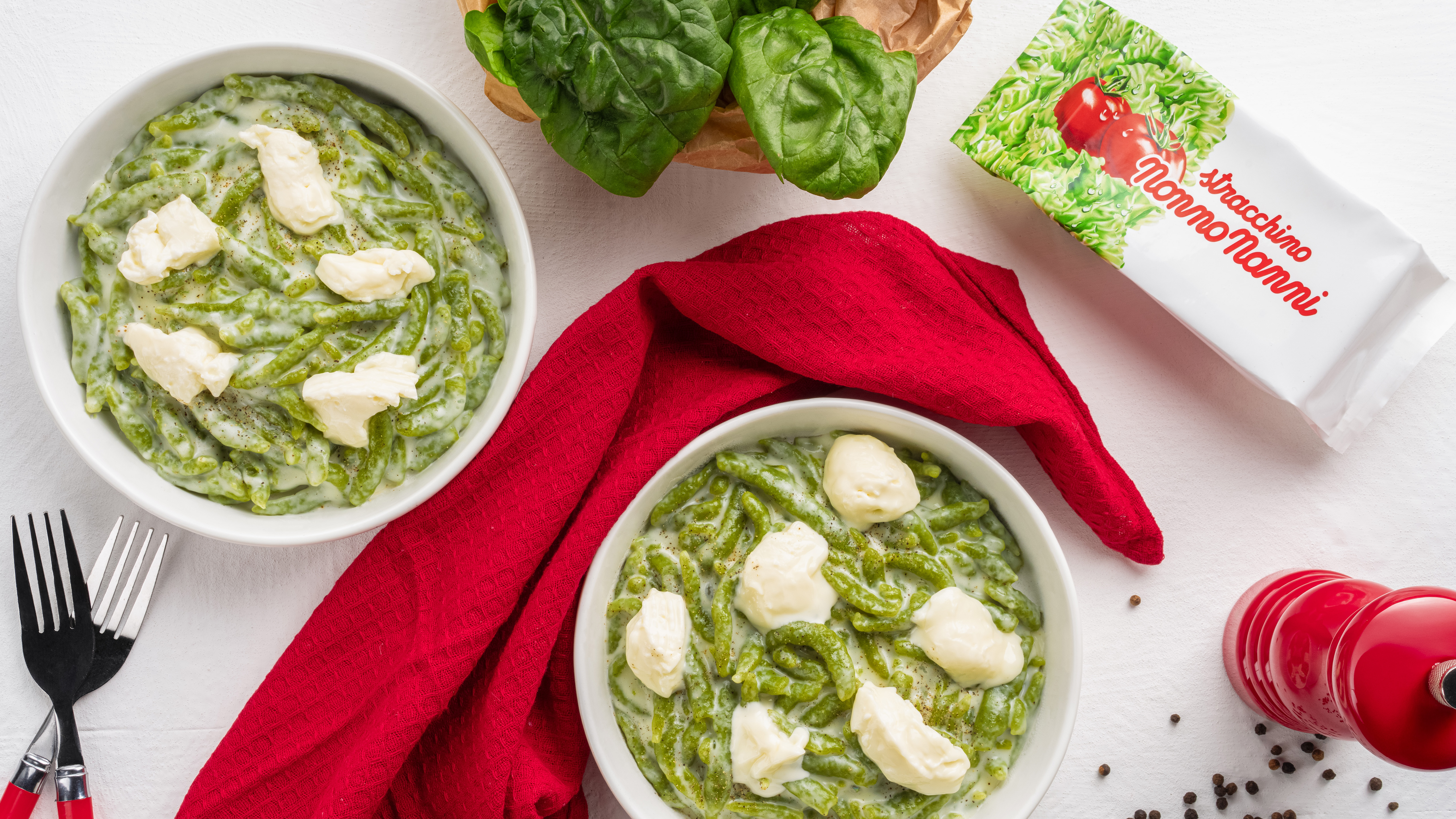 Due ciotole di pasta verde con crema e stracchino nonno nanni accanto a basilico tovagliolo rosso posate e macinapepe su sfondo bianco