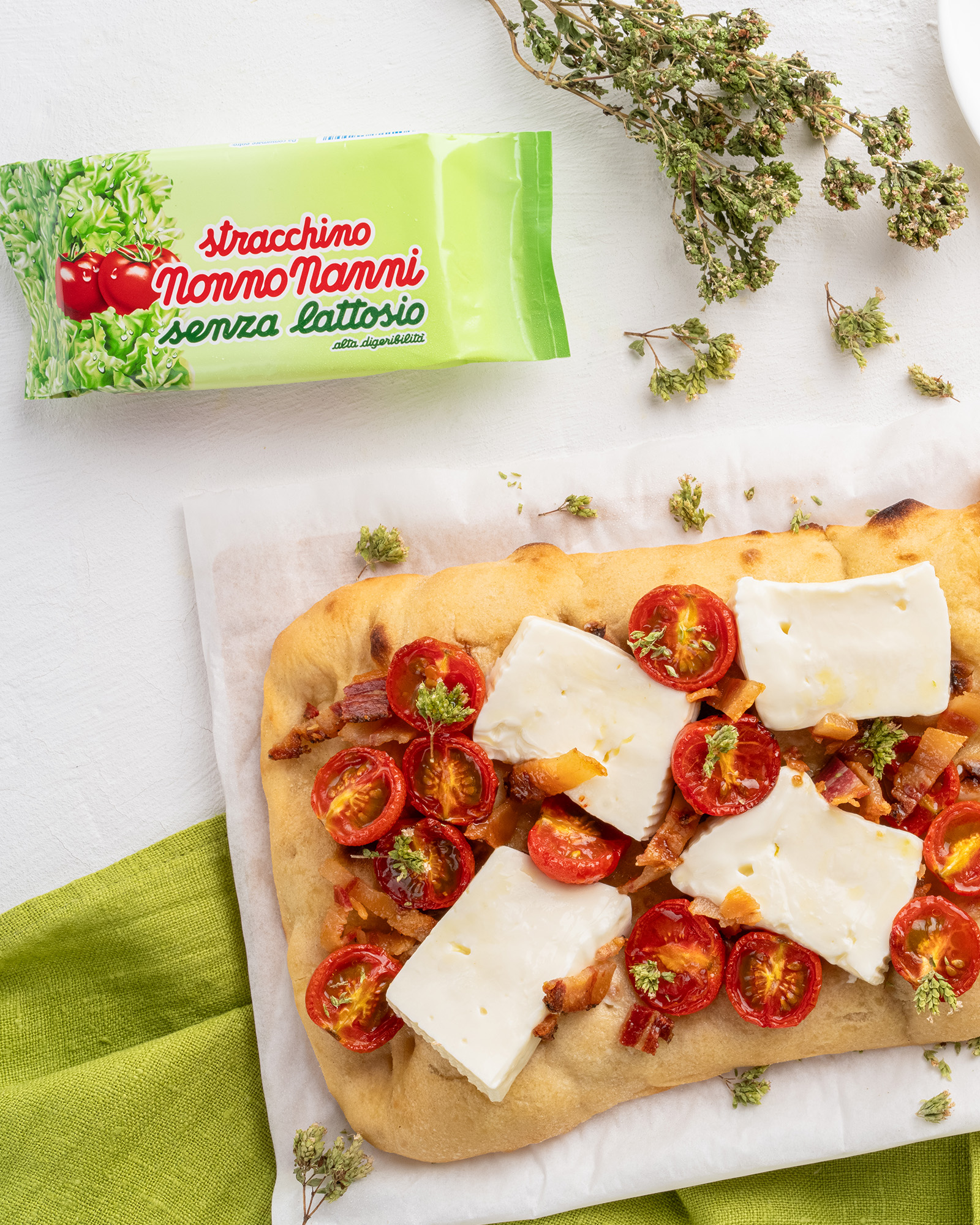 Focaccia con pomodorini stracchino e pancetta accanto a confezione di stracchino senza lattosio nonno nanni ed erbe aromatiche su sfondo bianco