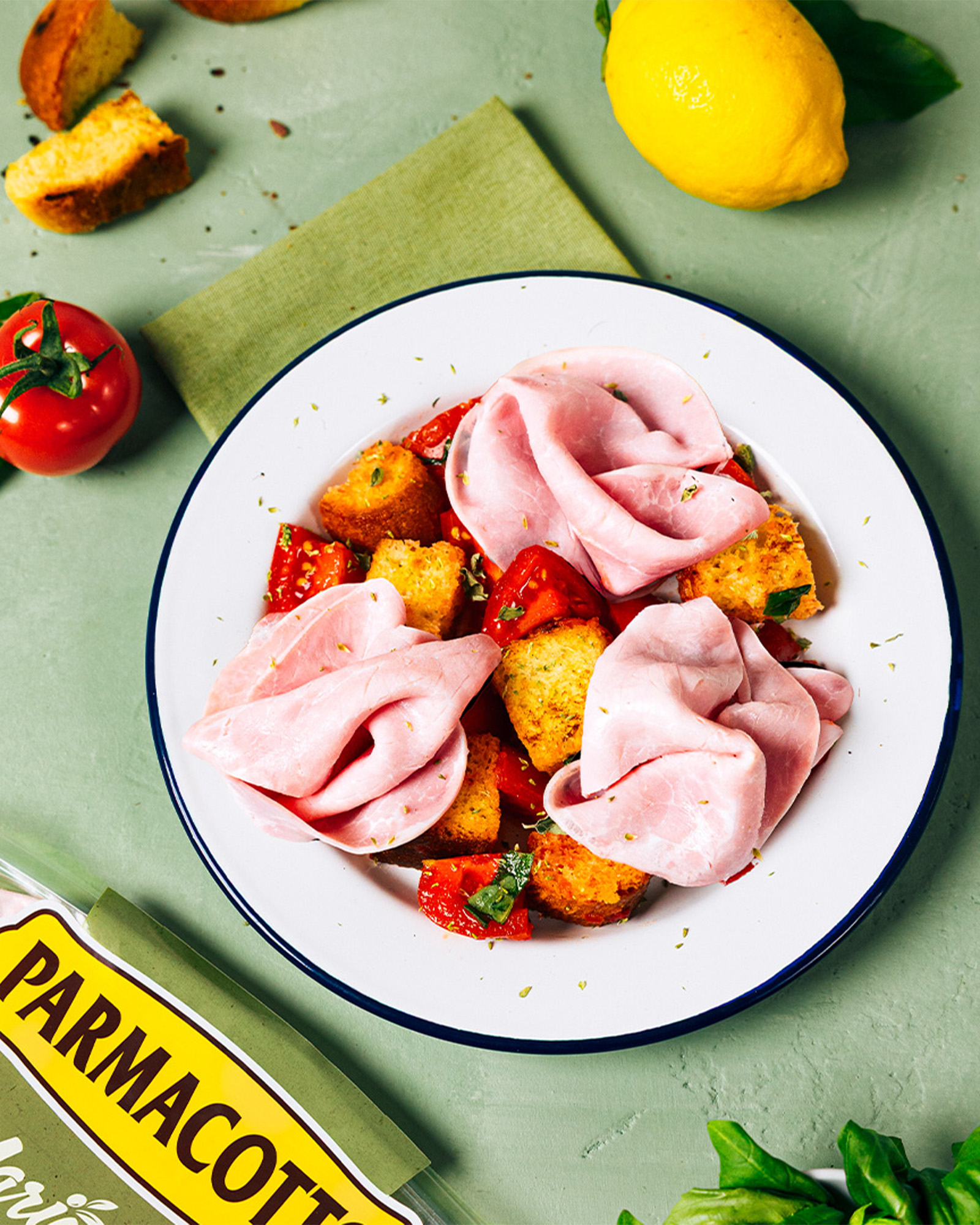 Piatto con mortadella crostini pomodorini ed erbe aromatiche su sfondo verde con limone pomodoro e confezione di parmacotto parzialmente visibile
