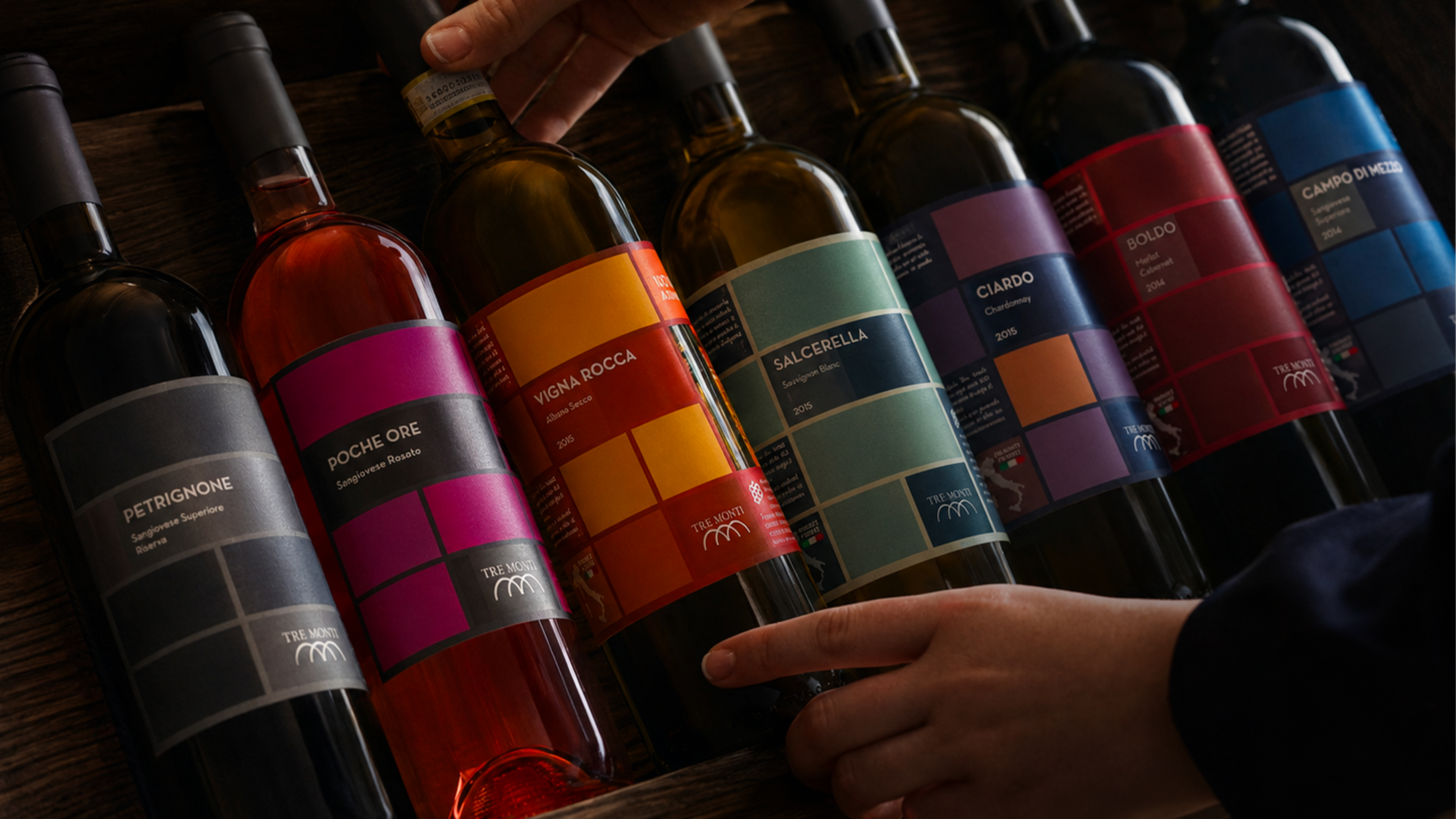 Bottiglie di vino tre monti con etichette colorate disposte su un piano in legno e selezionate da due mani